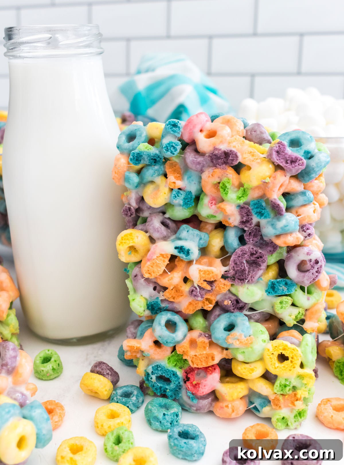 Closeup on a stack of three Froot Loops Marshmallow Treats sitting on a white table next to a glass of milk.