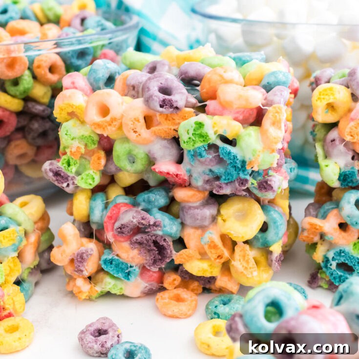 Close-up on a stack of two Froot Loops Marshmallow Treats sitting on a white table in front of glass bowls filled with Froot Loop Cereal and Mini Marshmallows.