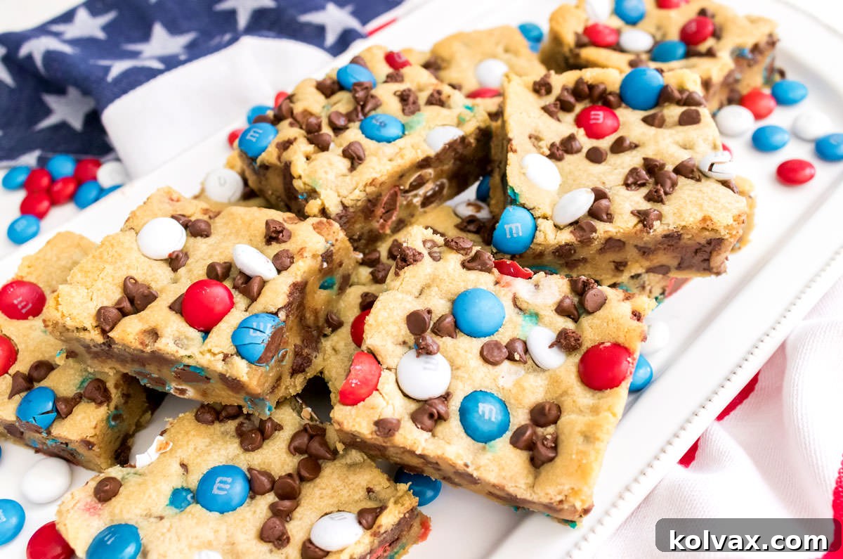 Closeup on a batch of Patriotic M&M Chocolate Chip Cookie Bars on a white dessert platter sitting on an American Flag Kitchen towel, showcasing their vibrant red, white, and blue colors and delicious texture.