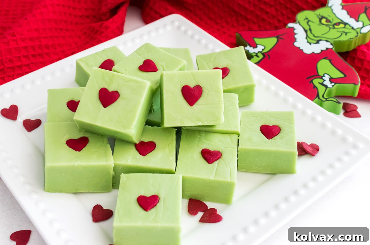 A close-up of a white dessert plate showcasing vibrant green Grinch Fudge pieces, each topped with a red heart sprinkle, surrounded by festive Grinch-themed holiday decorations.