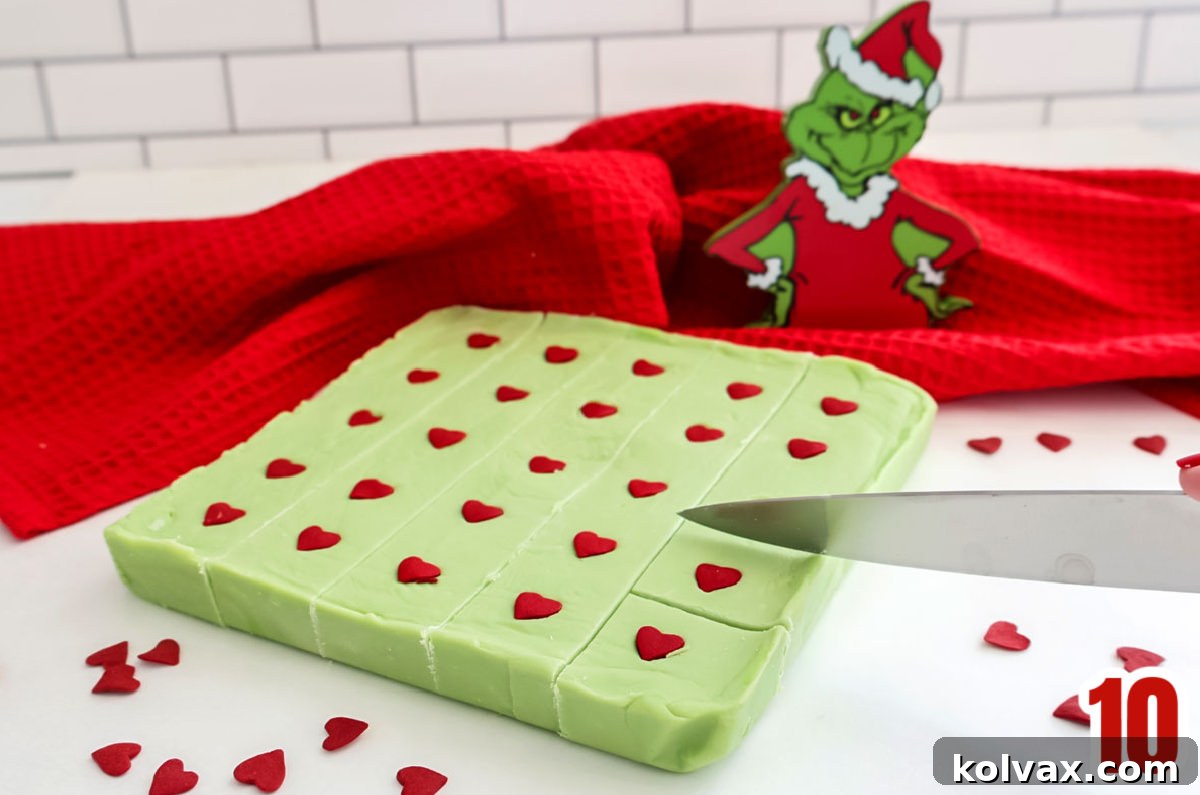A close-up shot of the vibrant green Grinch Fudge being carefully cut into individual, bite-sized pieces on a white cutting board, revealing the perfect consistency.