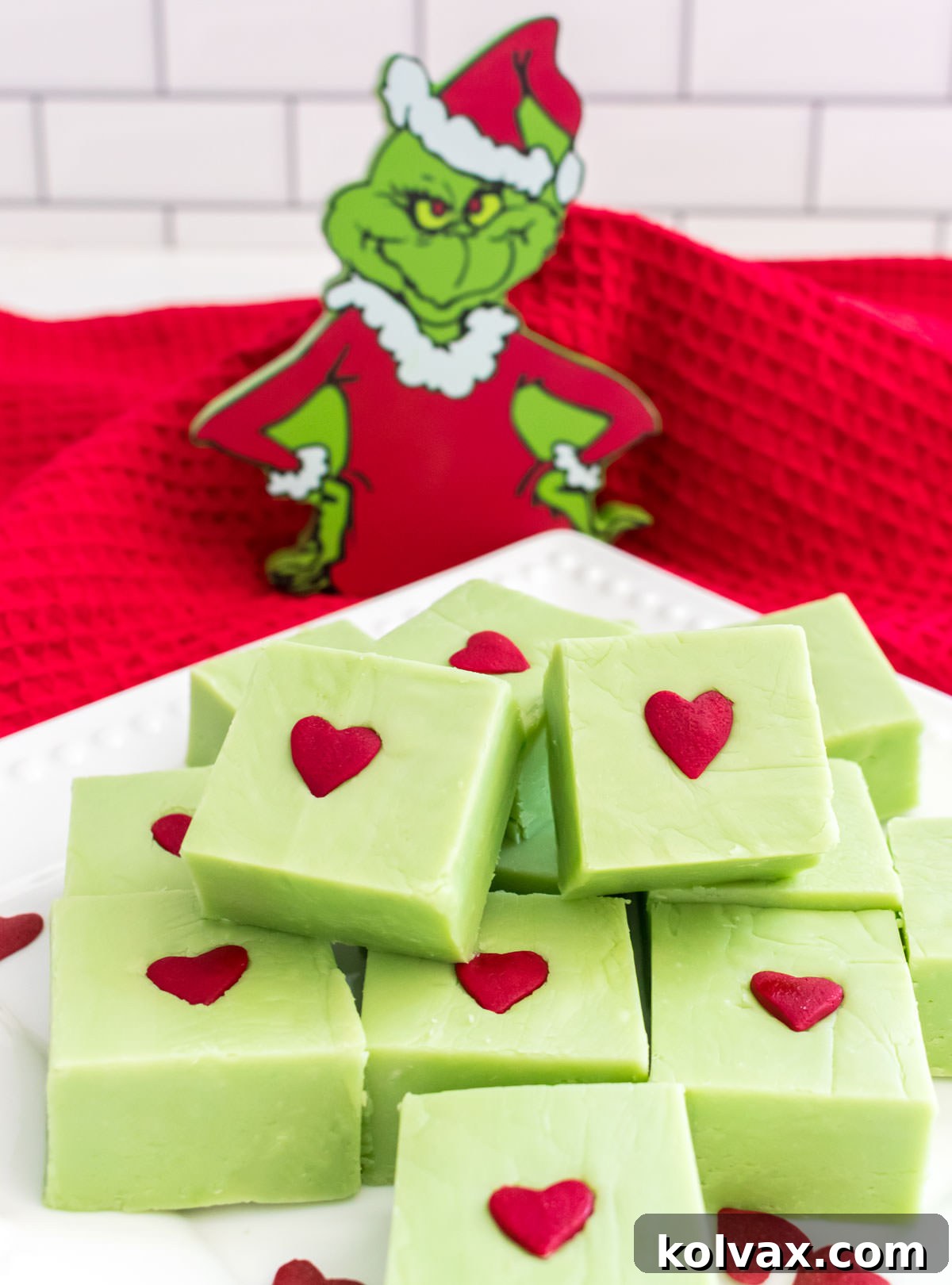A close-up of a white dessert plate abundantly filled with freshly cut Easy Grinch Fudge pieces, each adorned with a red heart, placed in front of a whimsical Grinch Christmas ornament.