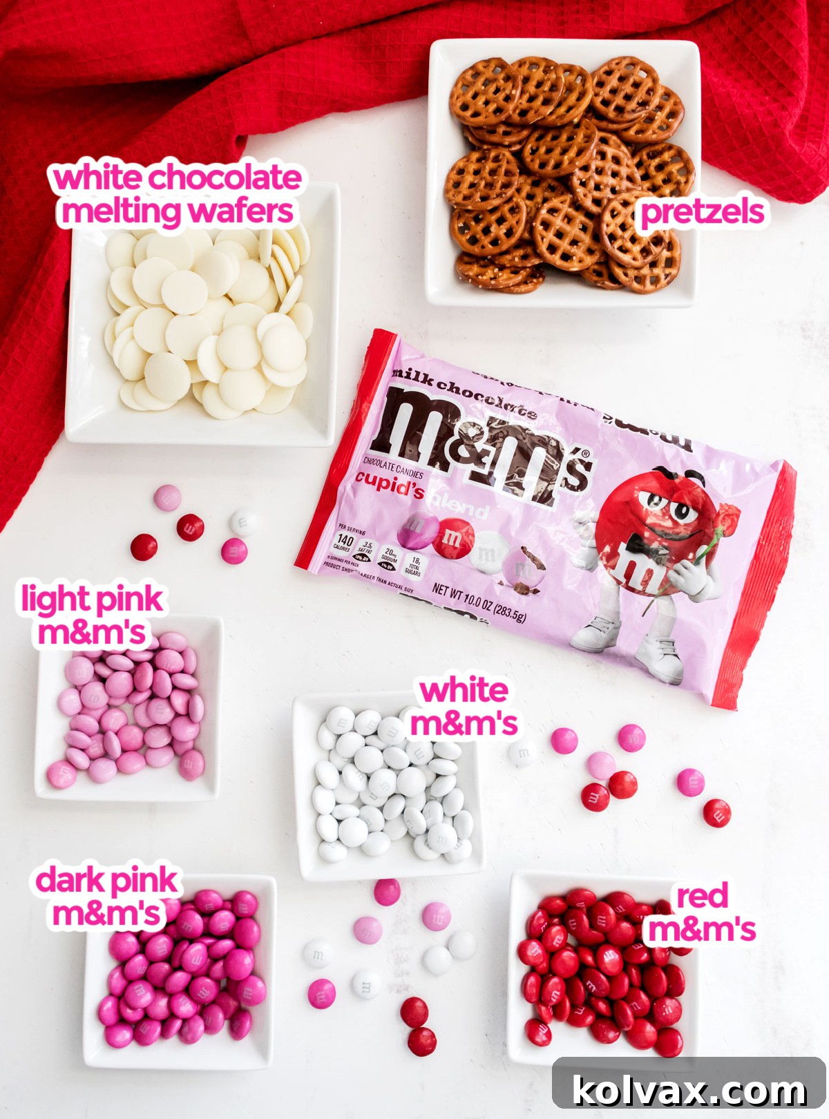 A flat lay photograph showcasing all essential ingredients for Rose Garden Flower Pretzel Bites: a bag of waffle pretzels, a bag of white chocolate candy melts, and bags of red, white, and pink M&M's candies.
