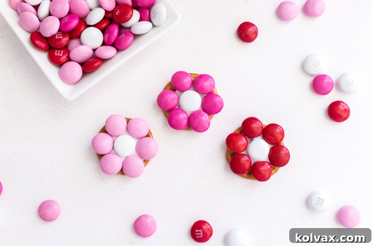 A close-up view of three completed Rose Garden Flower Pretzel Bites, featuring red, pink, and magenta petals with white centers, resting on a white surface surrounded by loose M&M's.