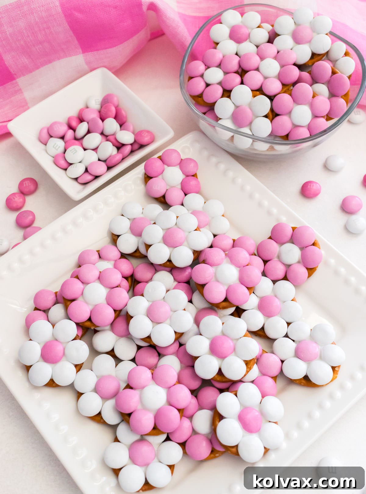 A elegant white dessert plate filled with Pretty in Pink Flower Pretzel Bites sits alongside a small glass bowl of the same treats, all resting on a white table with a cozy pink plaid kitchen towel draped nearby.