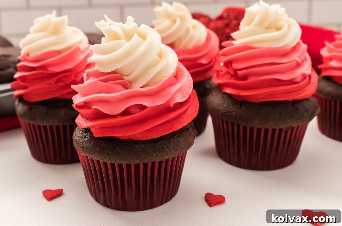 Close up of five Valentine's Day Cupcakes sitting on a white table surrounded by red heart sprinkles. Each cupcake features a beautiful three-color swirl of red, dark pink, and white buttercream frosting, topped with tiny red heart sprinkles.