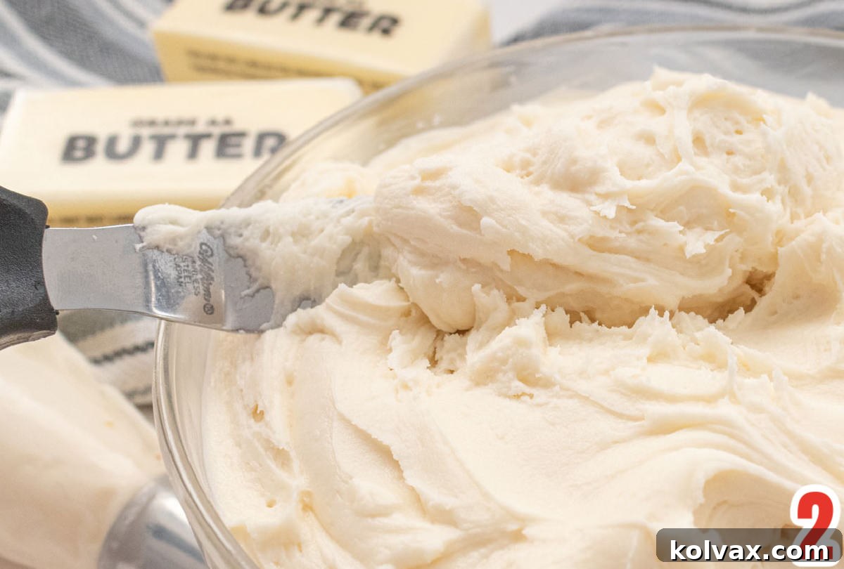 Close up on a glass bowl filled with freshly made Best Buttercream Frosting and an offset spatula, with two sticks of butter and a mixer in the soft-focus background.