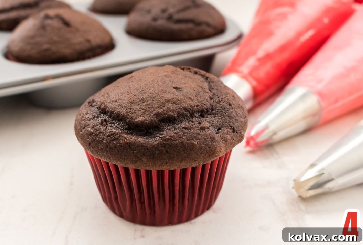 Close up of a chocolate cupcake in a red liner, with three distinct piping bags filled with red, dark pink, and white frosting ready for decoration.
