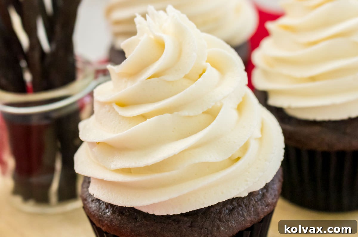 A perfectly piped cupcake topped with generous swirls of homemade Best Buttercream Frosting, sitting elegantly in front of other frosted cupcakes and a clear jar filled with fragrant vanilla beans, highlighting the key flavor ingredient.