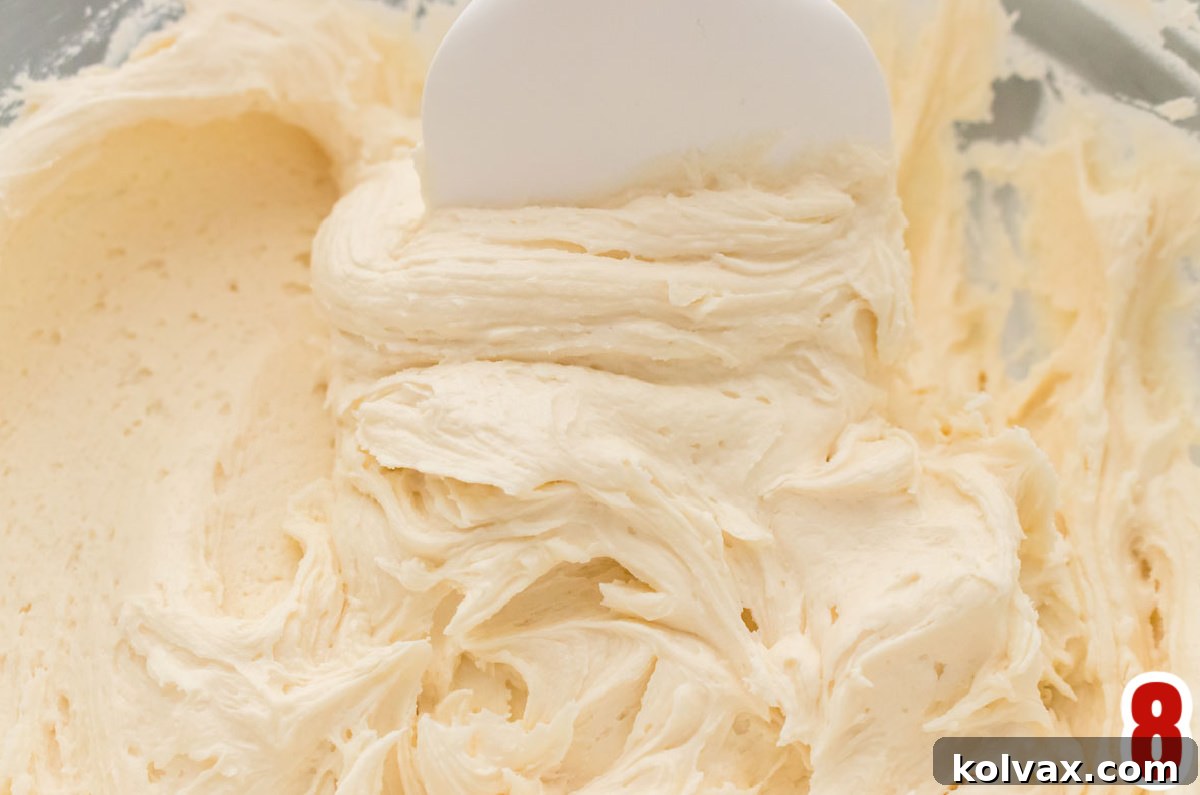 A close-up shot of a clear glass mixing bowl filled with the finished Best Buttercream Frosting, perfectly smooth, light, and fluffy, with a clean white rubber spatula resting inside the bowl, ready for tasting and use.