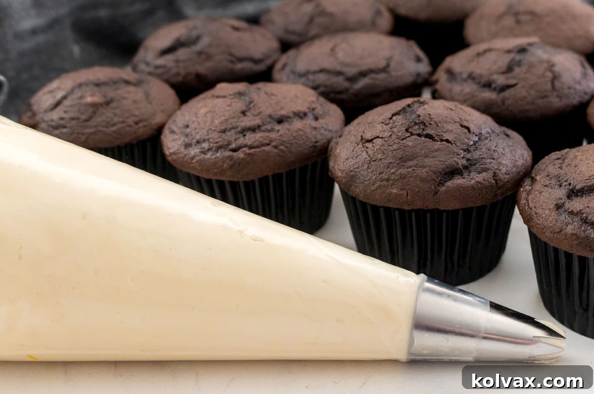 A pastry decorating bag brimming with luscious homemade buttercream frosting, ready to be piped onto a tray of unfrosted cupcakes in the background, illustrating the versatility of this recipe for decoration.