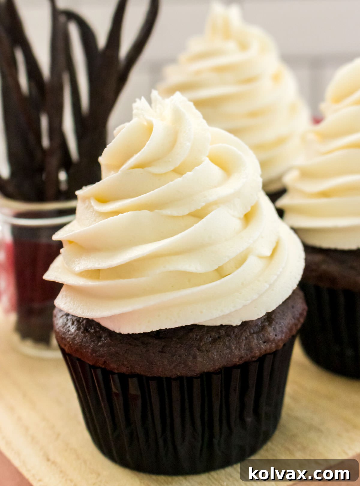 A rich chocolate cupcake, exquisitely topped with a generous swirl of The Best Buttercream Frosting and delicately garnished with fine chocolate shavings, rests invitingly on a rustic cutting board beside a charming jar of vanilla beans, emphasizing the frosting's versatile appeal.