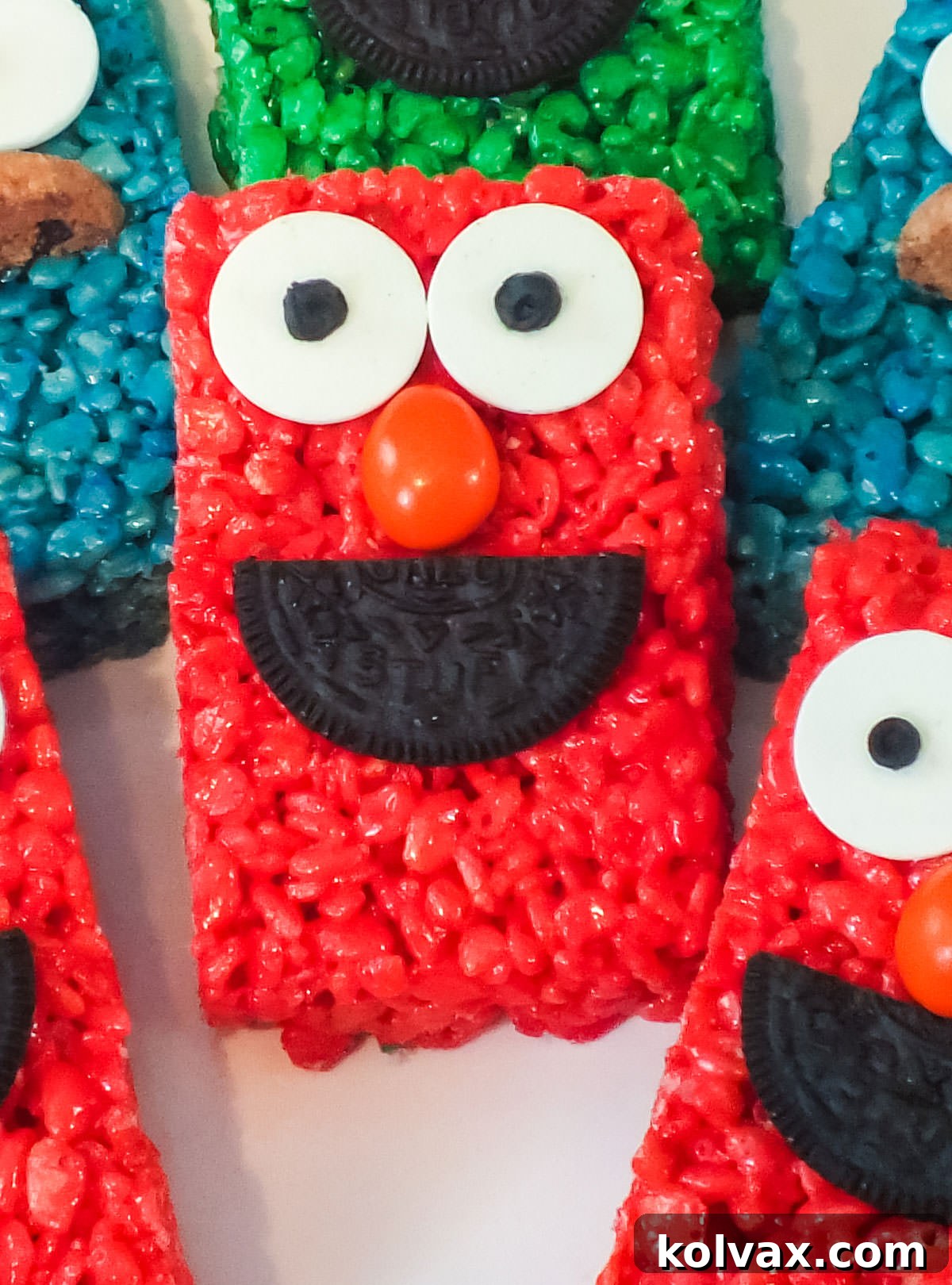 Closeup of a perfectly crafted Elmo Rice Krispie Treat, featuring his iconic red color, candy eyes, M&M nose, and Oreo mouth, resting on a white serving platter next to other colorful Sesame Street themed Rice Krispie Treats.