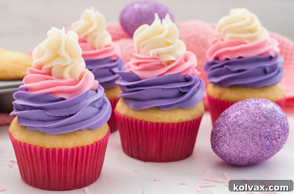 Close-up of four vibrantly frosted pastel Easter Cupcakes on a white surface, accompanied by a decorative Easter egg, a soft pink towel, and a baking pan filled with unadorned vanilla cupcakes in the background, showcasing the final product and baking process.