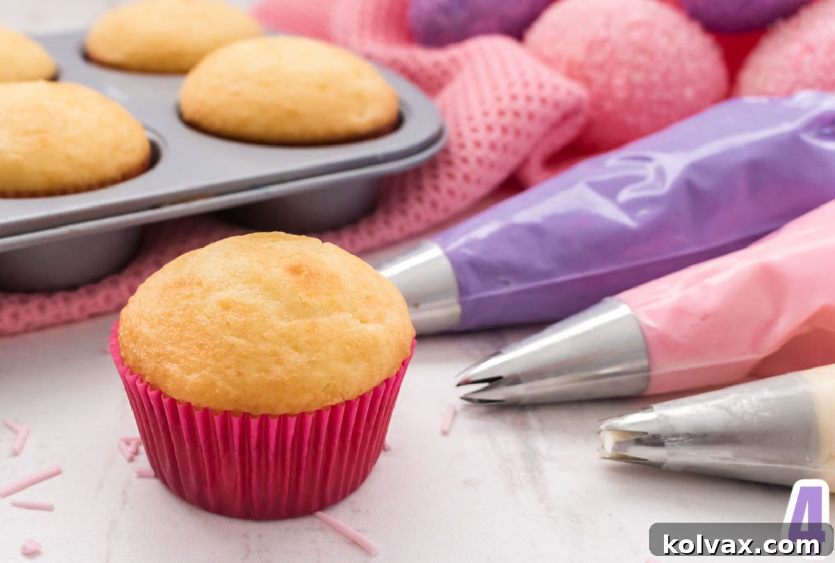 A close-up shot of a single vanilla cupcake nestled in a charming pink liner, positioned beside three separate piping bags filled with different colors of frosting and a baking pan holding more vanilla cupcakes, ready for the decorating process.