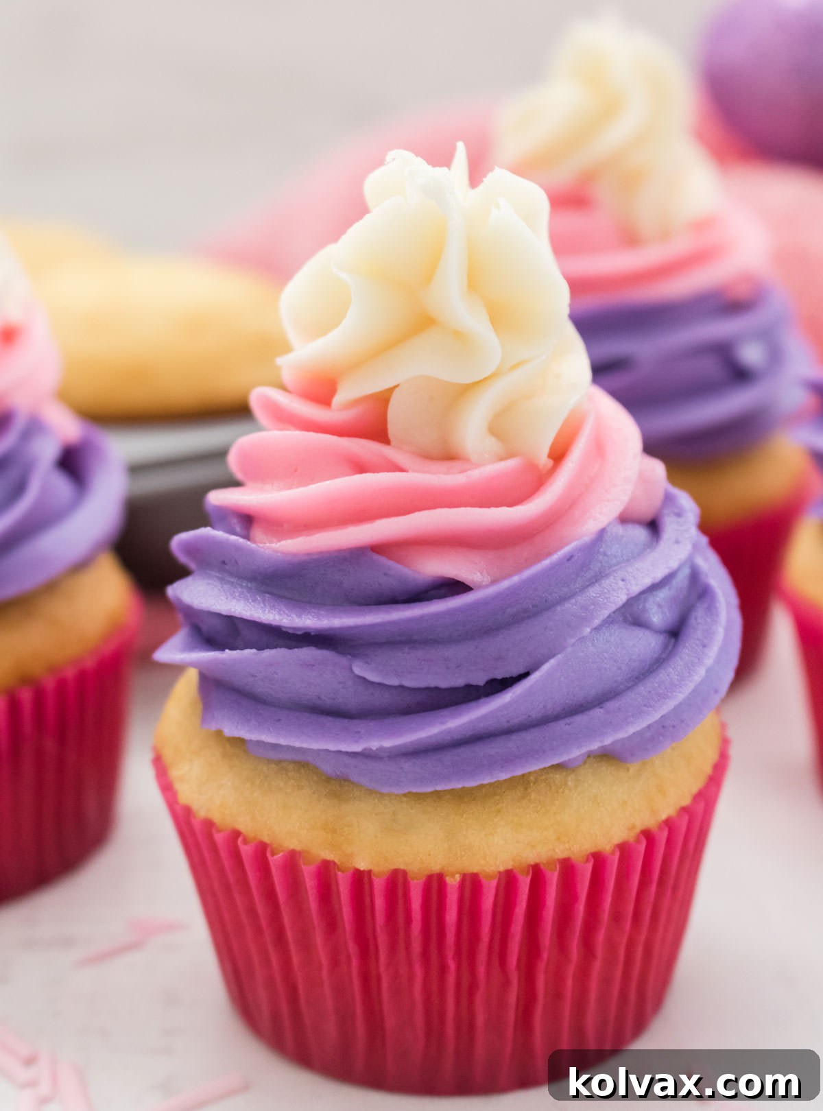 A captivating close-up of a single, perfectly piped Easter Cupcake, showcasing its intricate pastel frosting swirl, positioned on a white table. In the soft-focus background, more frosted cupcakes, a baking tin, and a pink table linen create an inviting spring scene.