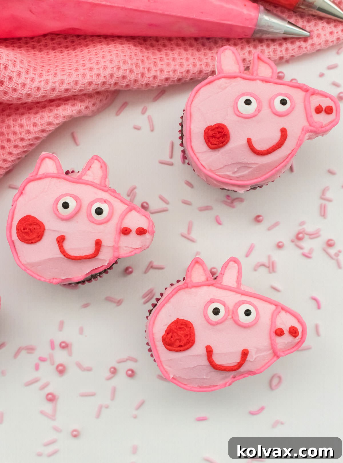 A beautiful closeup of three finished Peppa Pig Cupcakes, artfully arranged on a pristine white surface, adorned with cheerful pink sprinkles, and set against a backdrop of a soft pink table linen and a bowl of complementary pink frosting.