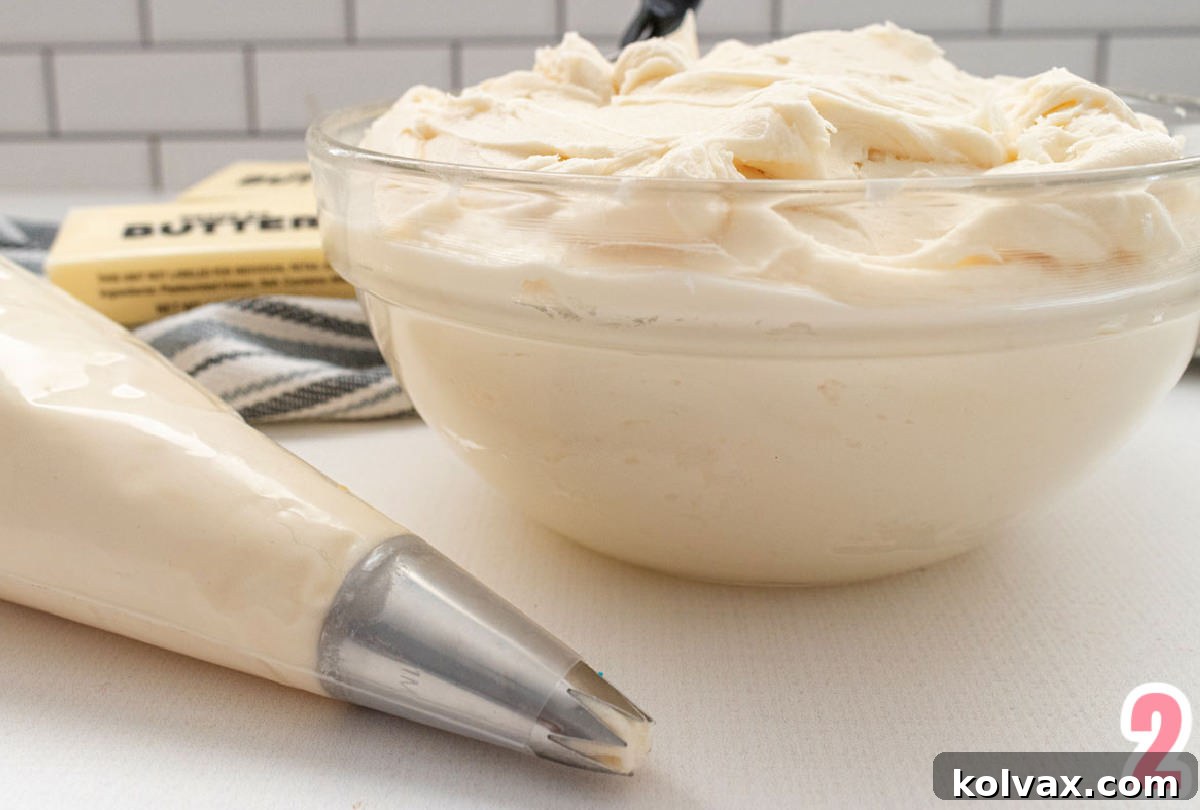 A clear glass bowl brimming with light, fluffy homemade buttercream frosting, positioned beside a piping bag filled with the same frosting, ready for vibrant coloring and decoration.