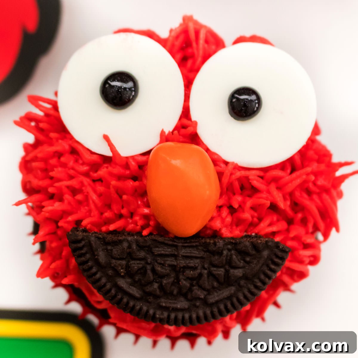 Closeup of an Elmo Cupcake sitting on a white table. A bright red Elmo cupcake with large white candy eyes and an orange candy melt nose, radiating cheerfulness.