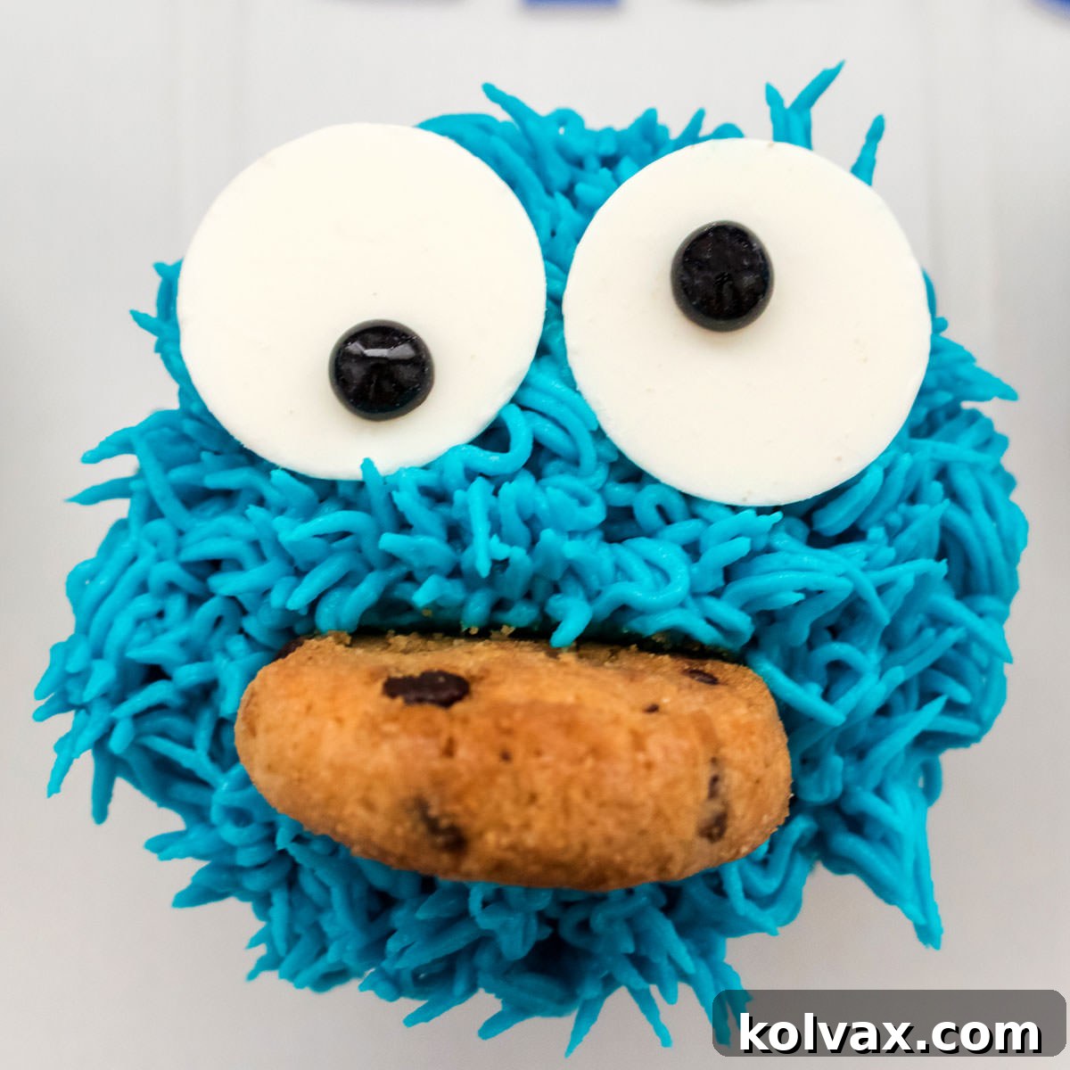 Closeup of a Cookie Monster Cupcake sitting on a white table. A vibrant blue Cookie Monster cupcake with large candy eyes, a wide mouth, and a piece of chocolate chip cookie stuck within it.