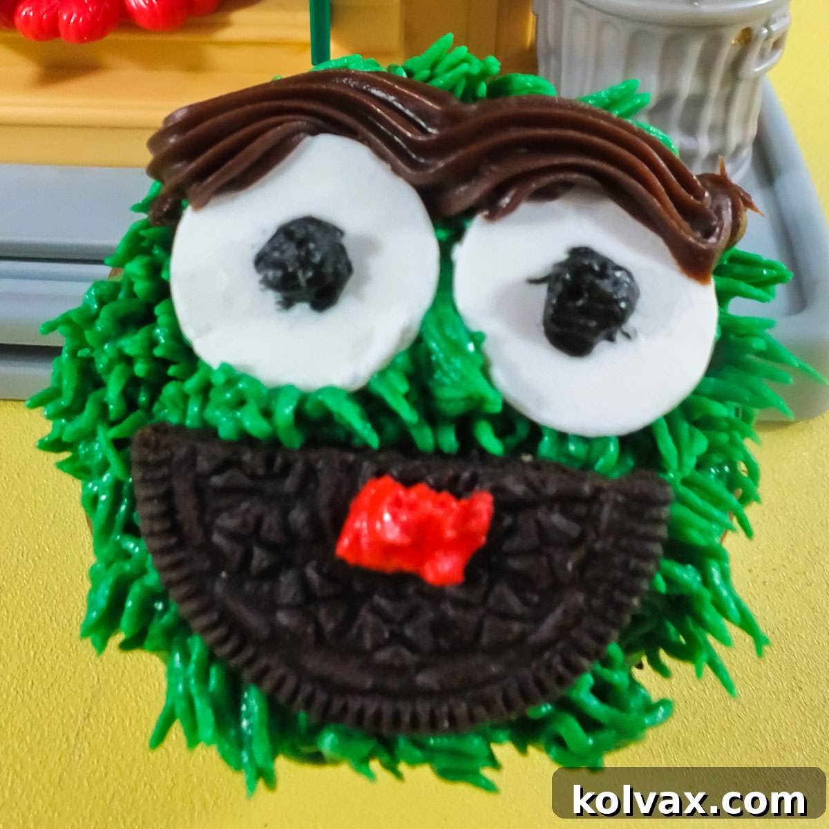 Closeup of an Oscar the Grouch Cupcake sitting on a yellow surface in front of a Sesame Street toy. A green Oscar the Grouch cupcake with grumpy white candy eyes, a wide Oreo mouth, a distinct chocolate unibrow, and a small red tongue.
