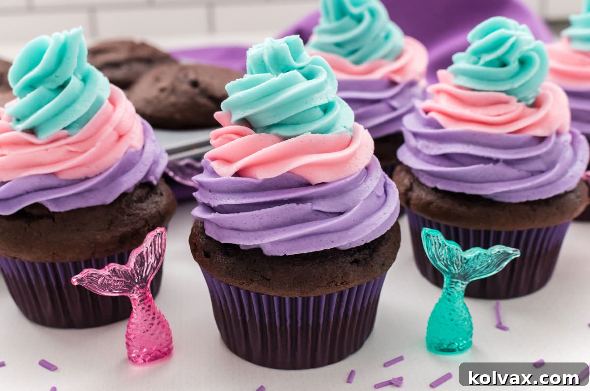Closeup on four Mermaid Cupcakes sitting on a white table in front of a tin filled with cupcakes and a purple table linen.