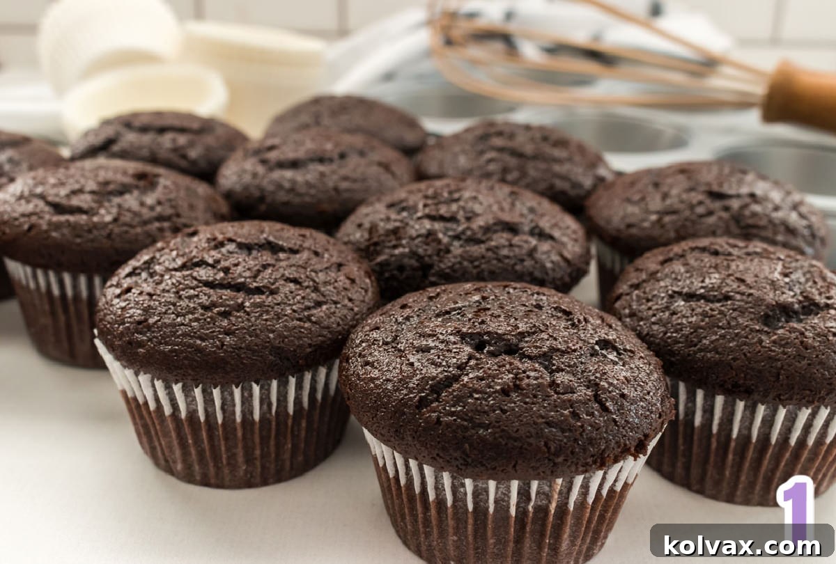Ten chocolate cupcakes sitting on a white table in front of a cupcake tin and white cupcake liners.