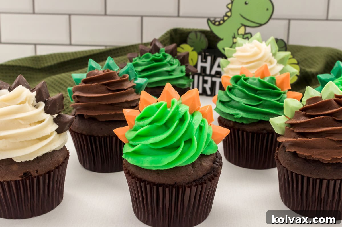 Closeup on eight vibrant Dinosaur Cupcakes with Stegosaurus spikes, resting on a white table with a green kitchen towel and a cheerful cartoon dinosaur in the background.