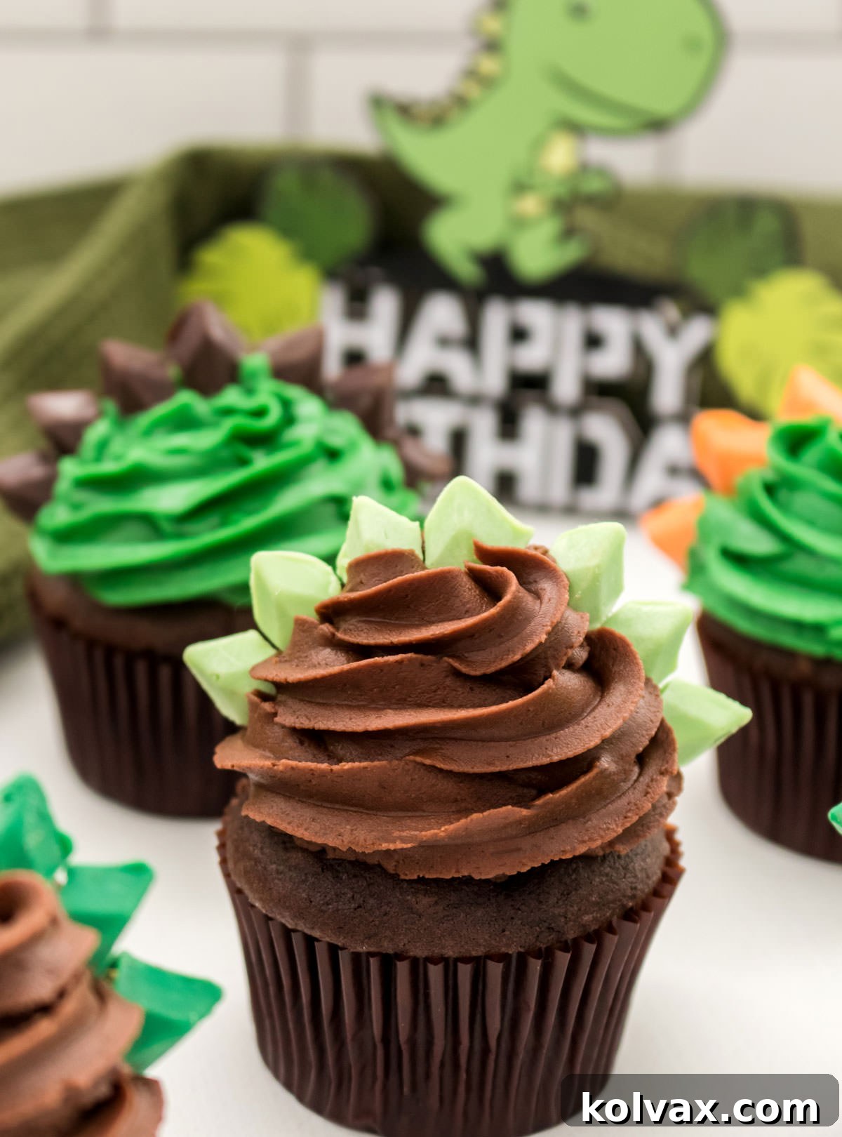 A close-up shot of a single Dinosaur Cupcake, featuring rich chocolate frosting and striking green Stegosaurus spikes, with other colorful cupcakes softly blurred in the background.