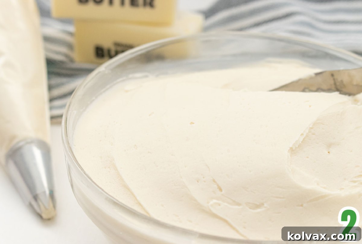 A clear glass bowl brimming with light and fluffy buttercream frosting, positioned beside a stick of butter and a piping bag filled with the same delicious frosting, awaiting decoration.
