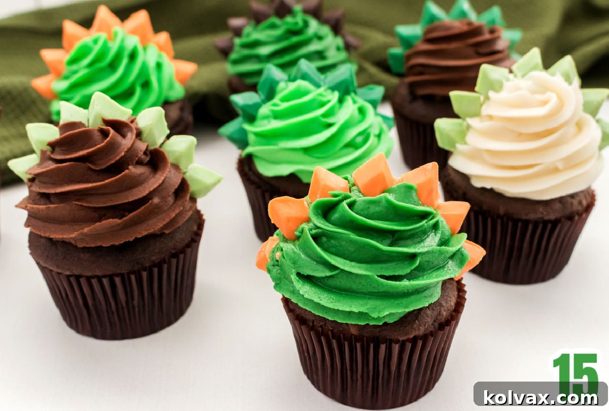 A close-up of seven beautifully decorated Dinosaur Cupcakes, each featuring colorful frosting and distinct Stegosaurus spikes, arranged on a white table in front of a green kitchen towel.