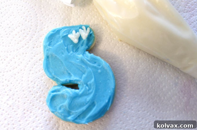 Decorating frosted sugar cookies with white buttercream dinosaur footprints, adding a themed touch.
