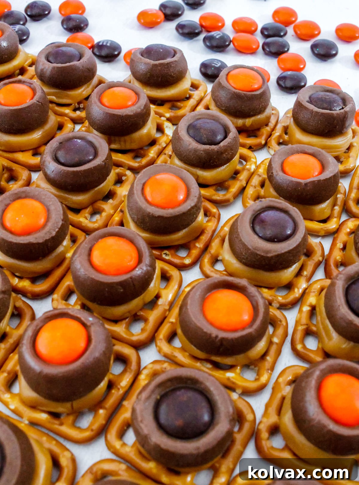 Spooky Salty Sweet Pretzel Delights 7 A mouth-watering close-up of a batch of Halloween Chocolate Caramel Pretzel Bites, resting on a white table, adorned with scattered orange and black M&M's, emphasizing their festive appeal.
