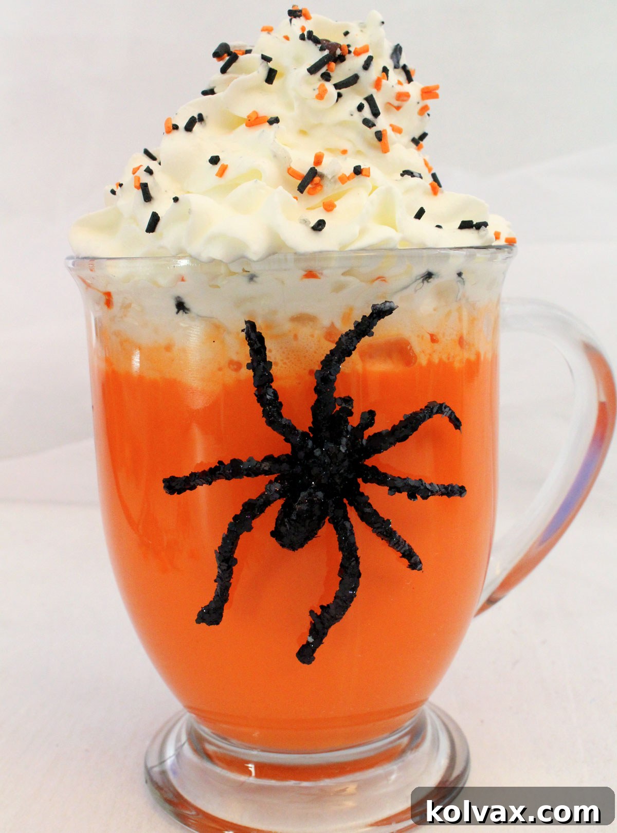 Closeup on a glass of Halloween Hot Vanilla Milk sitting on a white table with a toy spider on the glass.