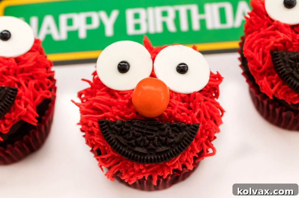 Close on three Elmo Cupcakes sitting on a white surface in front of a Happy Birthday Sesame Street Sign.