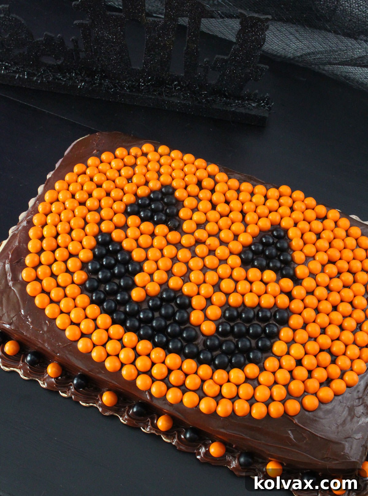 Closeup of a finished Easy Halloween Cake, a chocolate sheet cake adorned with a detailed Jack O' Lantern made from orange and black Sixlets, presented on a dark surface.