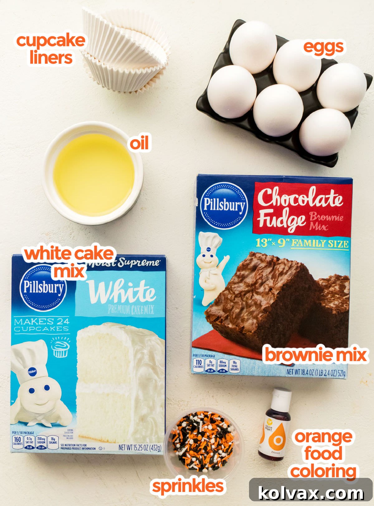 All the ingredients you will need to make Halloween Brownie Cupcakes including White Cake Mix, Brownie Mix, Eggs, Oil, Food Coloring and Halloween Sprinkles arranged on a white surface.