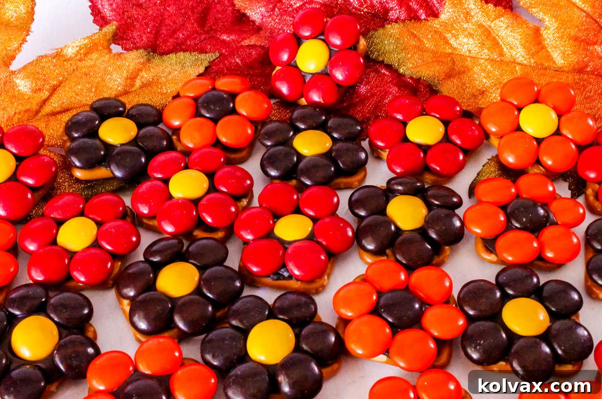Closeup on a batch of vibrant Harvest Flower Pretzel Bites laying on a white table next to Fall leaves decorations, showcasing their appealing colors and intricate M&M flower design.
