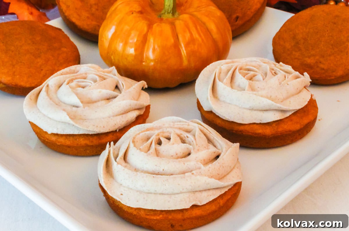 Closeup of three perfectly frosted Pumpkin Mini Cakes arranged on a pristine white platter, with a miniature decorative pumpkin subtly blurred in the background, evoking a sense of fall charm.