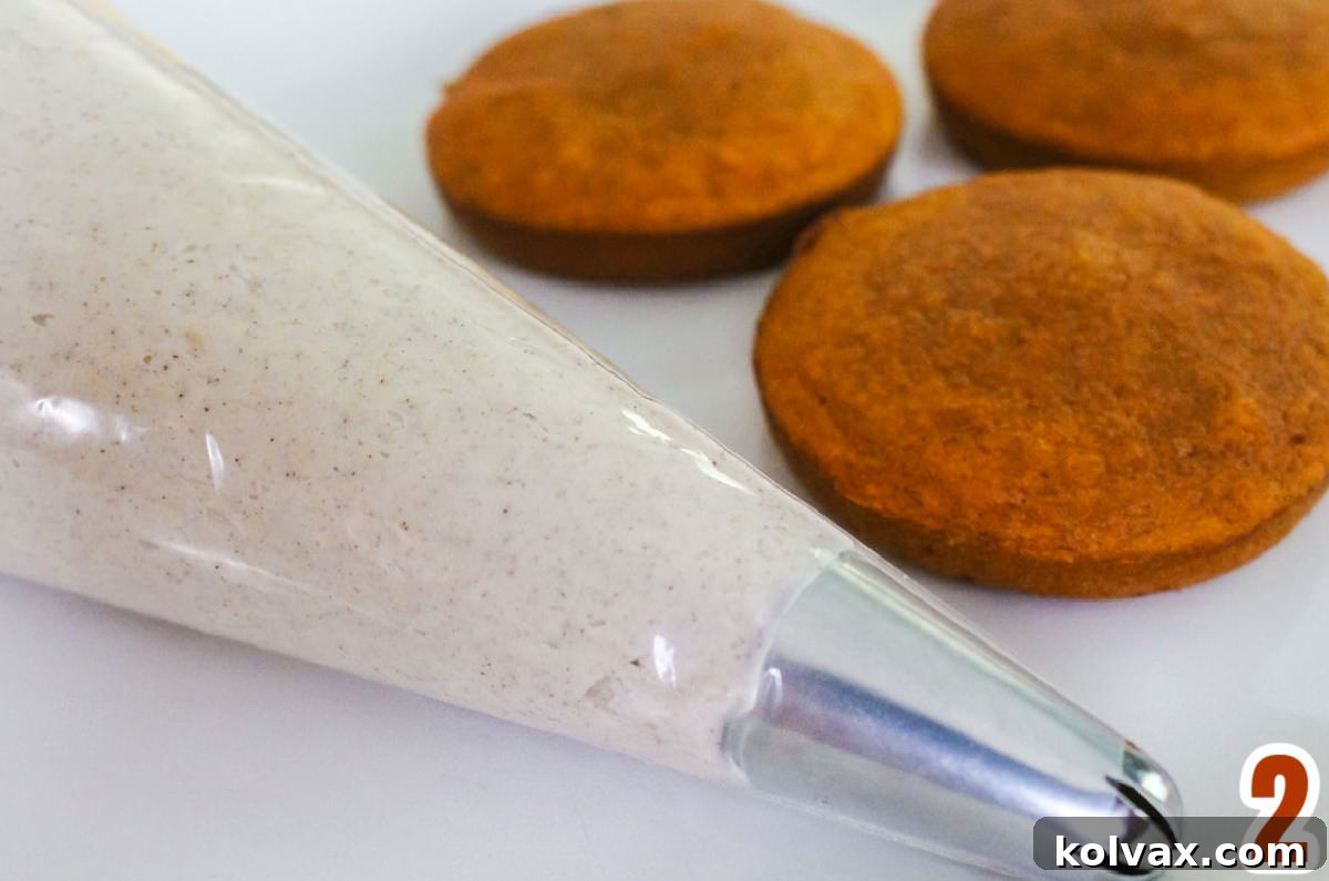 A clear piping bag, expertly filled with creamy cinnamon buttercream frosting, rests beside three unfrosted pumpkin mini cakes, awaiting their decadent topping, all on a crisp white surface.