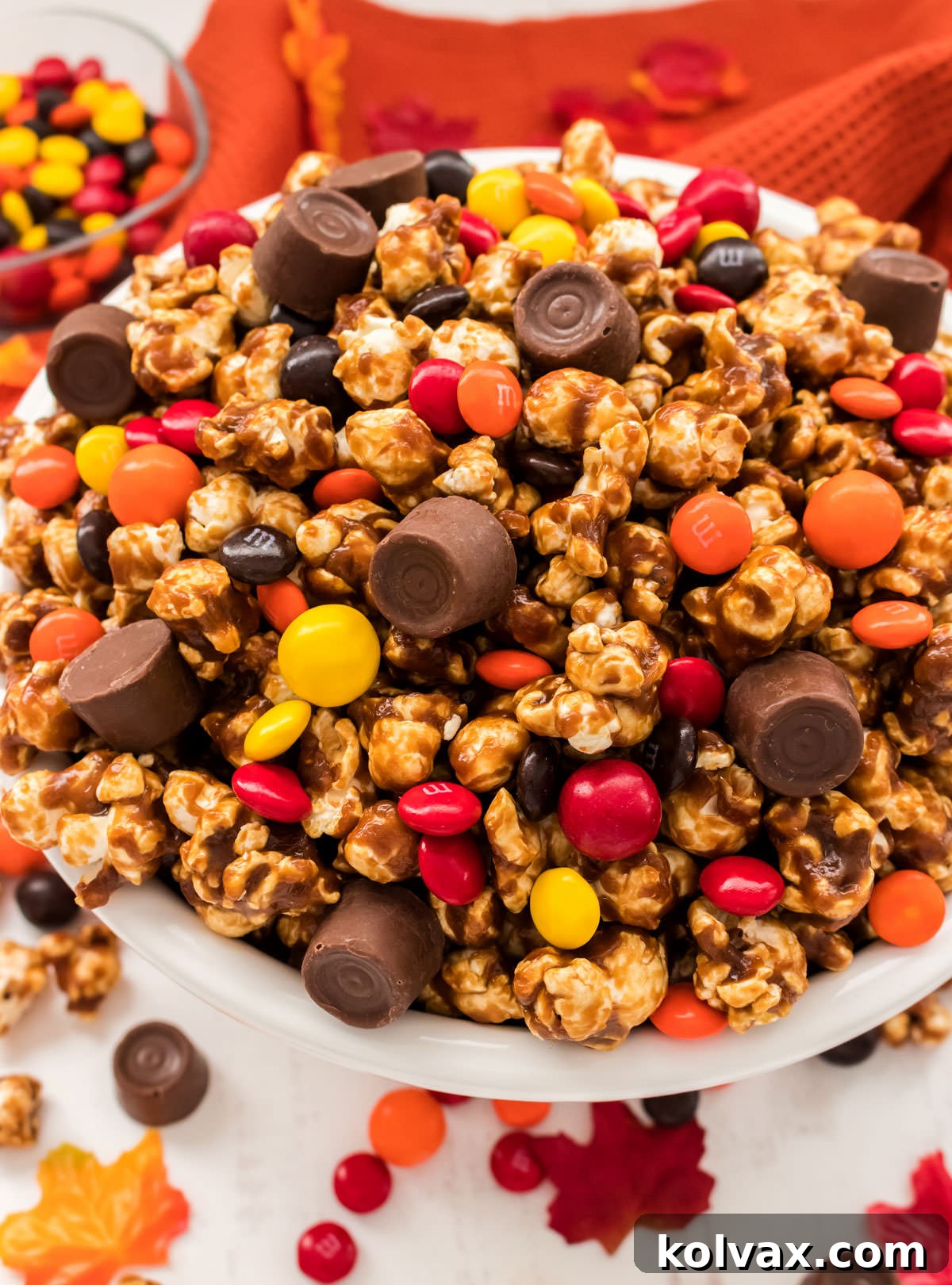 Closeup on a white serving bowl filled with Harvest Caramel Corn and sitting on a white table surrounded by Fall candy and autumn leaves.