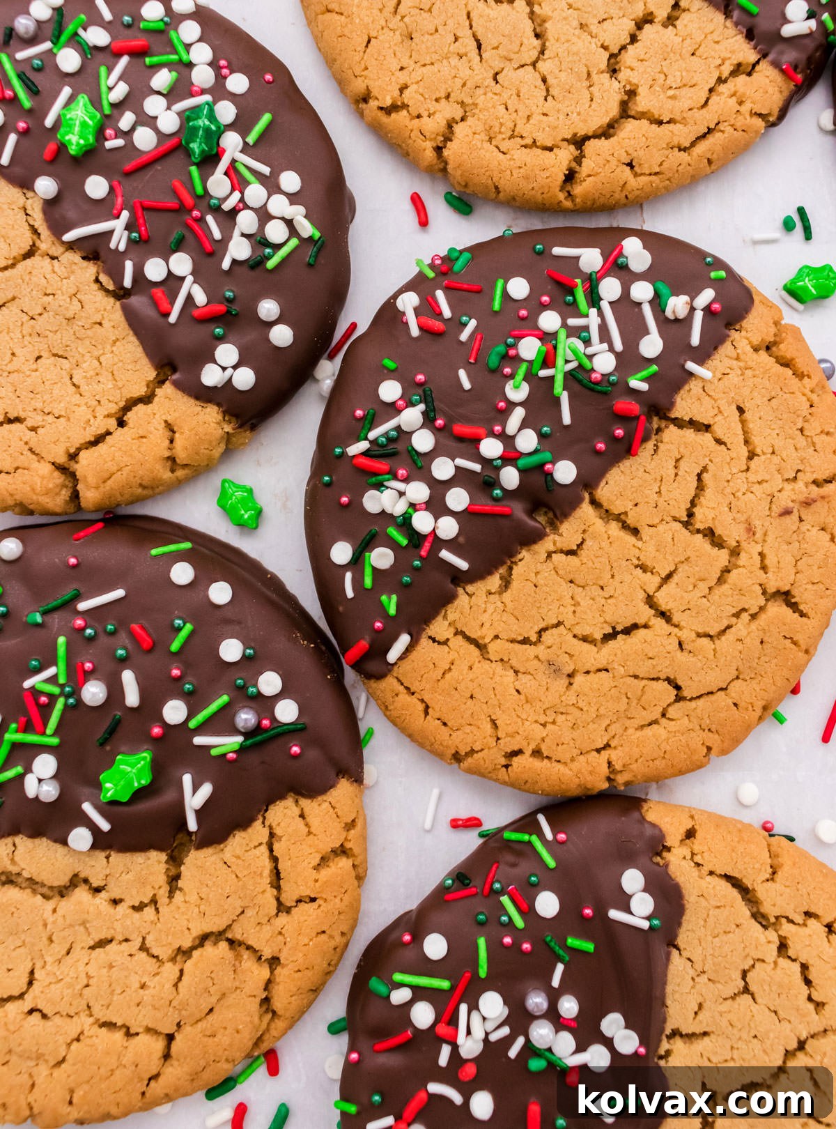 A close-up shot of five exquisitely decorated Chocolate Dipped Peanut Butter Cookies, lying flat on white parchment paper and artfully surrounded by colorful festive sprinkles, highlighting their holiday appeal.