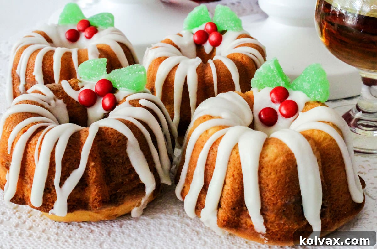 Four festive Christmas Mini Bundt Cakes, beautifully iced and decorated with holly berries, resting on a pristine white surface beside a glass of amber sherry, symbolizing holiday cheer.