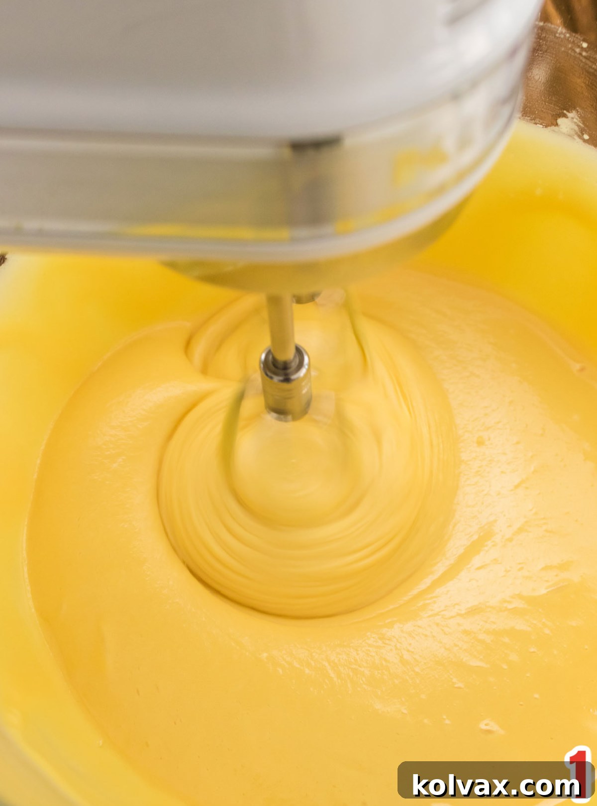 A close-up shot of the creamy cake batter being mixed with an electric hand mixer in a large glass bowl, showcasing the smooth, golden texture.
