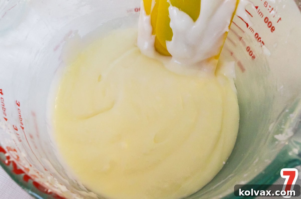 A clear glass measuring cup overflowing with smooth, glistening white cake icing, with a rubber spatula resting inside, indicating a perfect consistency.