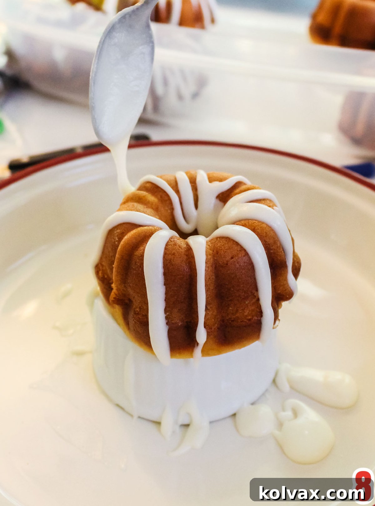 A close-up of a Mini Bundt Cake on a white surface, being artfully drizzled with sweet white icing from a spoon, creating an elegant cascade.