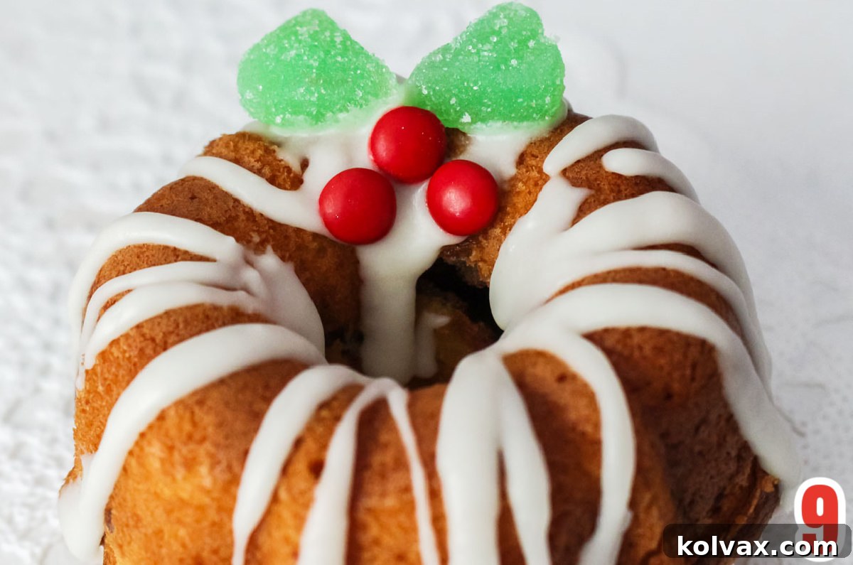 A perfectly iced Christmas Mini Bundt Cake adorned with a charming candy holly berry decoration made from red M&M's and green jelly slices, highlighting festive detail.