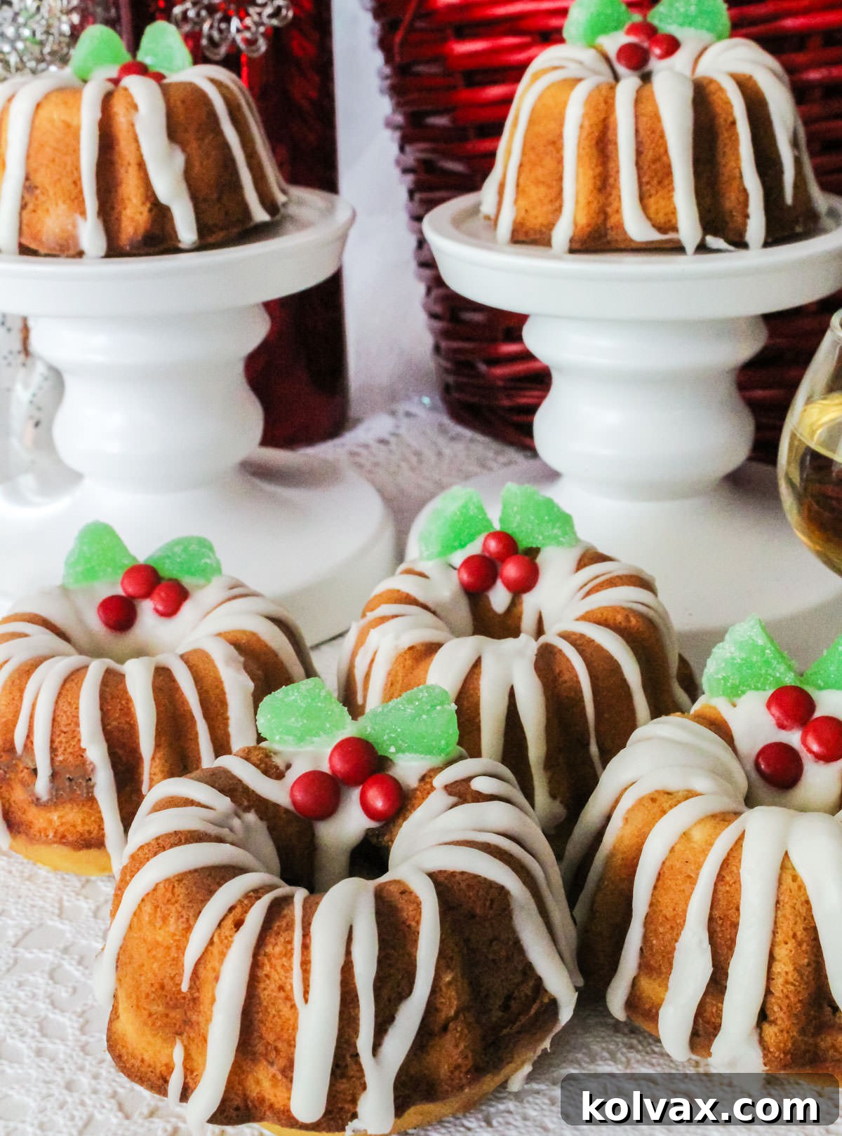 A beautifully arranged spread of six glistening Christmas Mini Bundt Cakes, each perfectly iced and adorned with festive holly berry decorations, presented elegantly on a white table.