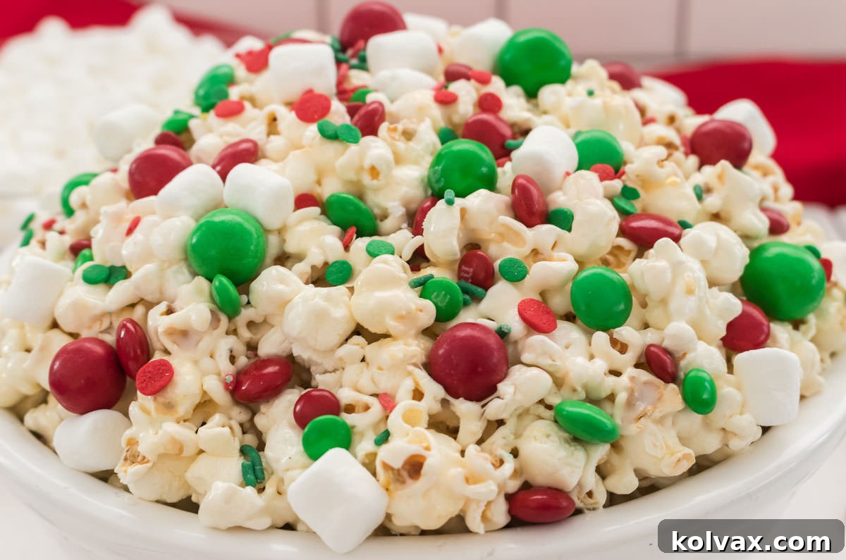 Closeup on a white serving bowl filled with Santa Crunch Popcorn sitting on a white table in front of a bowl of marshmallows and a red towel.