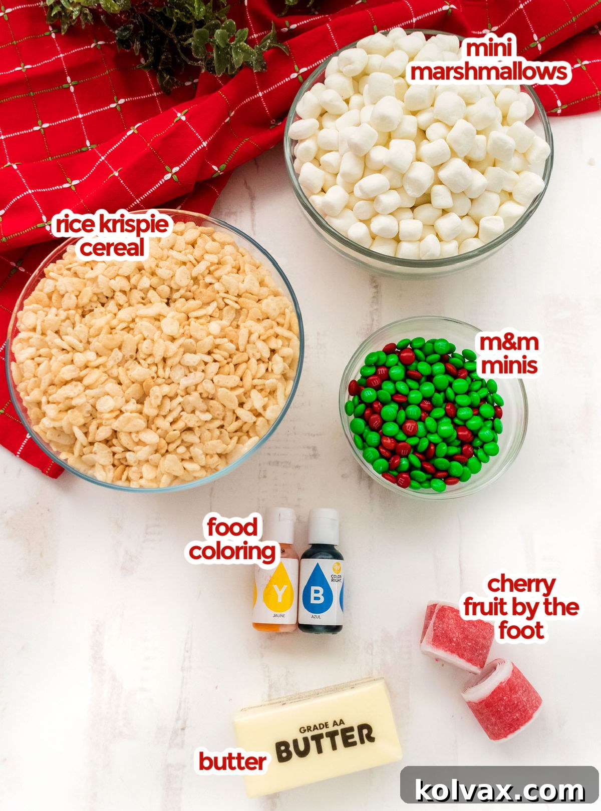 All the essential ingredients for Christmas Wreath Rice Krispie Treats laid out on a white surface: Mini Marshmallows, Rice Krispies Cereal, Mini M&M's, Green and Yellow Food Coloring, Butter, and Cherry Fruit by the Foot.